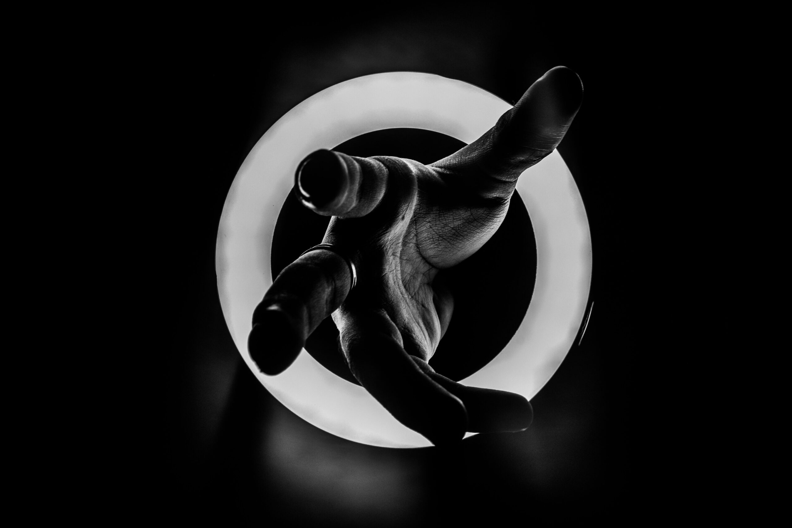 A dramatic black and white shot of a hand reaching through a glowing ring light, emphasizing shadows. The act of making something physical